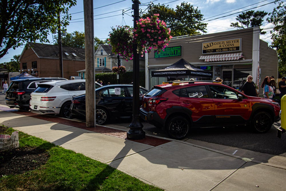 Cars parked on a street next to shops on a sunny day, with people walking by.