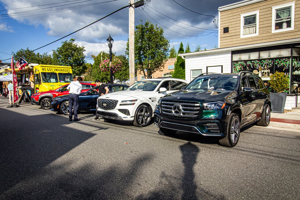 A busy outdoor street fair with various people looking at cars, a yellow 'We Got Empanadas!'' food truck, and buildings.