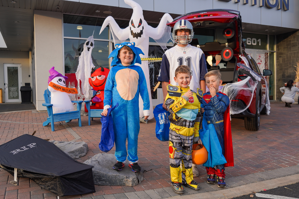 Four boys in Halloween costumes, with inflatable ghosts and a red car decorated for 'trunk or treat' in the background