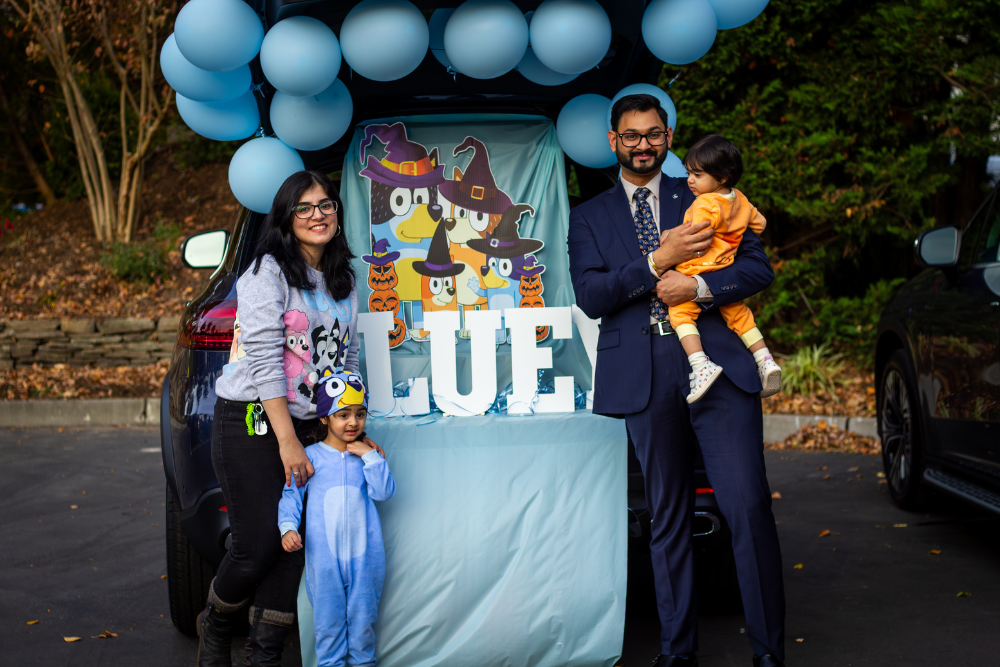 A family of four, including two young children dressed for Halloween, stands by a car decorated with Bluey characters.