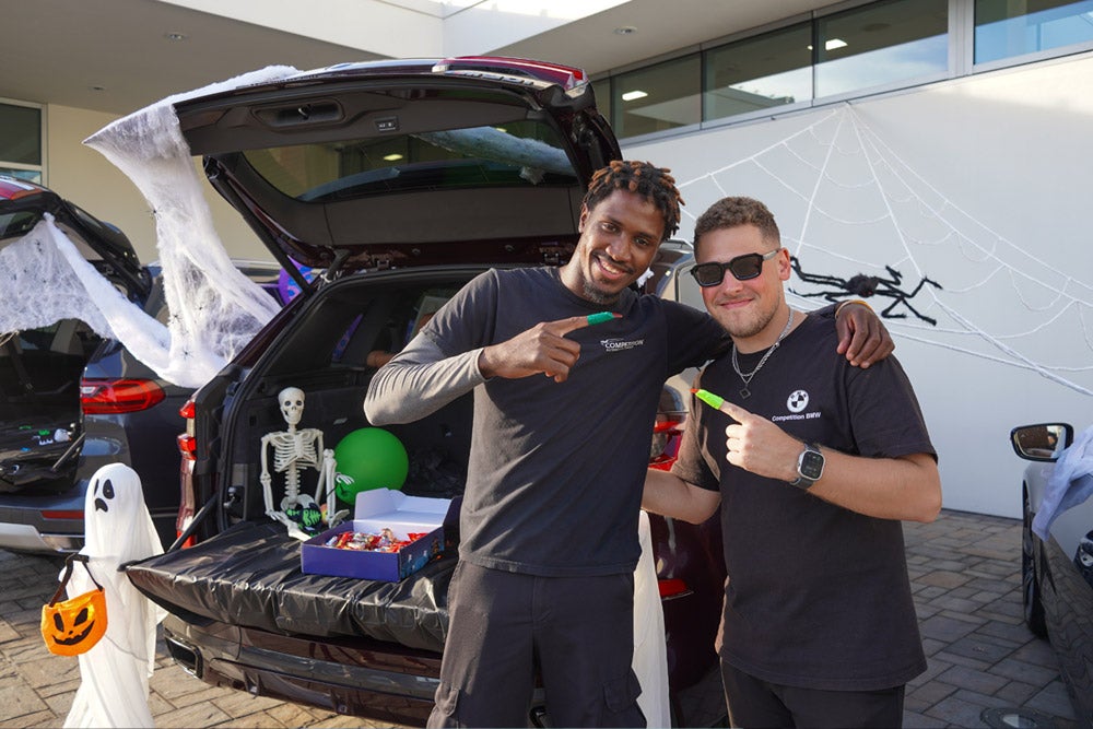 Two men smiling, one with dreadlocks and one in sunglasses, stand by a car trunk decorated for Halloween with a skeleton and candy.