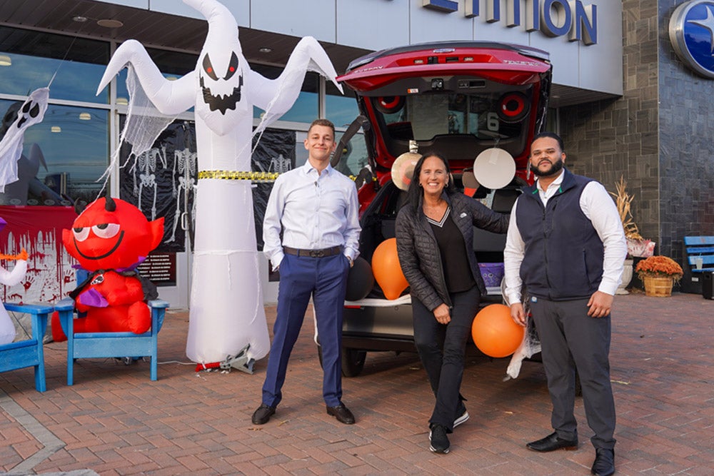 Three adults stand in front of a Halloween-decorated car dealership with inflatable ghosts and a red devil.