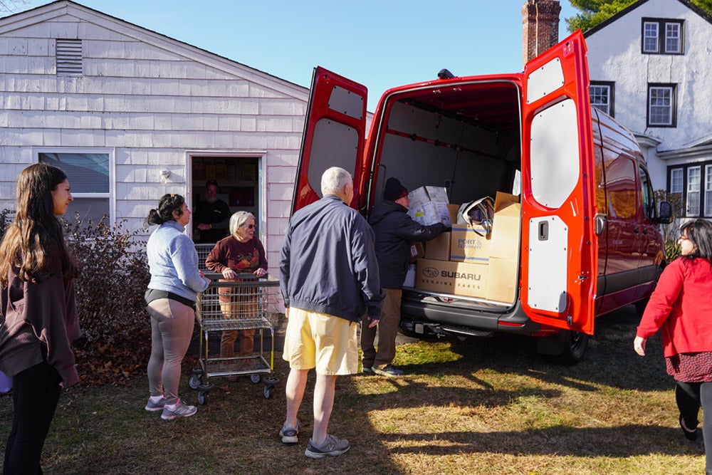 People load cardboard boxes from a red delivery van into a white house with a woman standing by a shopping cart.