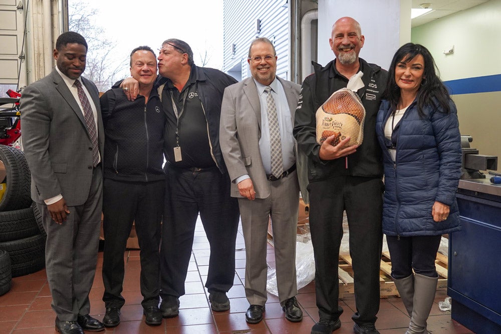 Six individuals, four men and two women, pose for a photo. A man on the right holds a netted brown ham.