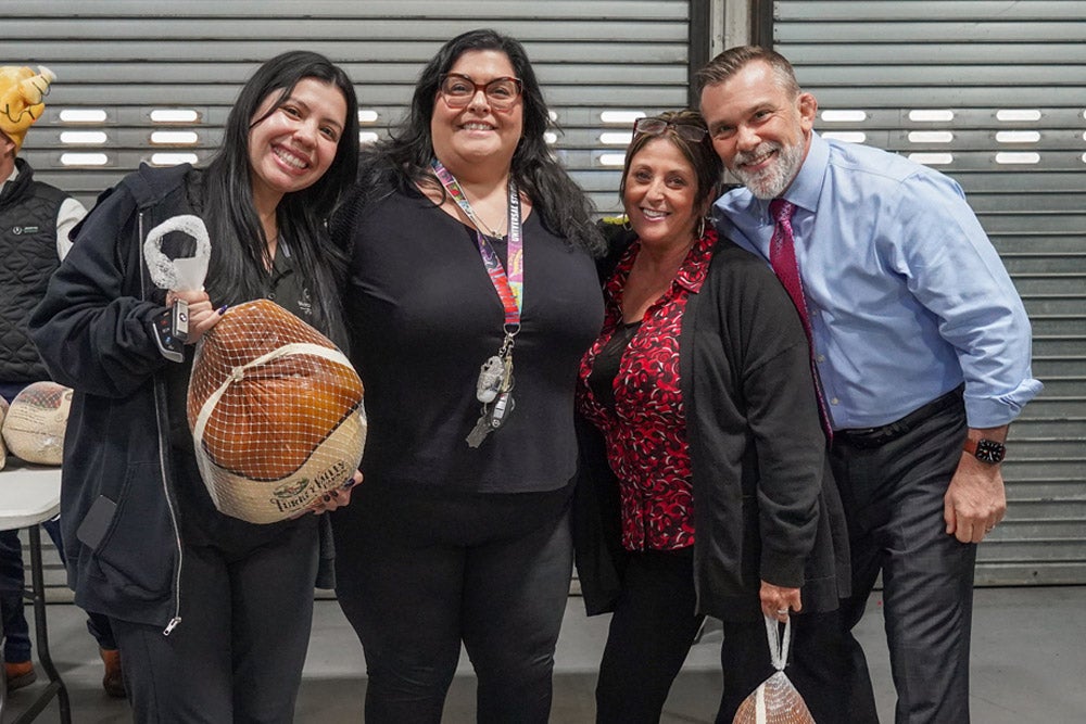 Four smiling people, two women holding turkeys, stand together against a corrugated metal wall.