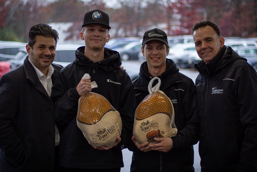 Four men, two holding turkeys, smiling for a photo, with cars in the blurry background.