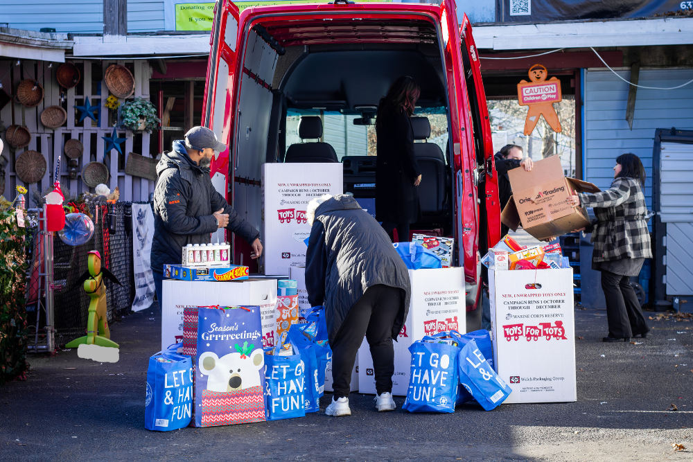 People loading gift bags and boxes of toys, including 'Toys for Tots' donations, into and around an open red van.