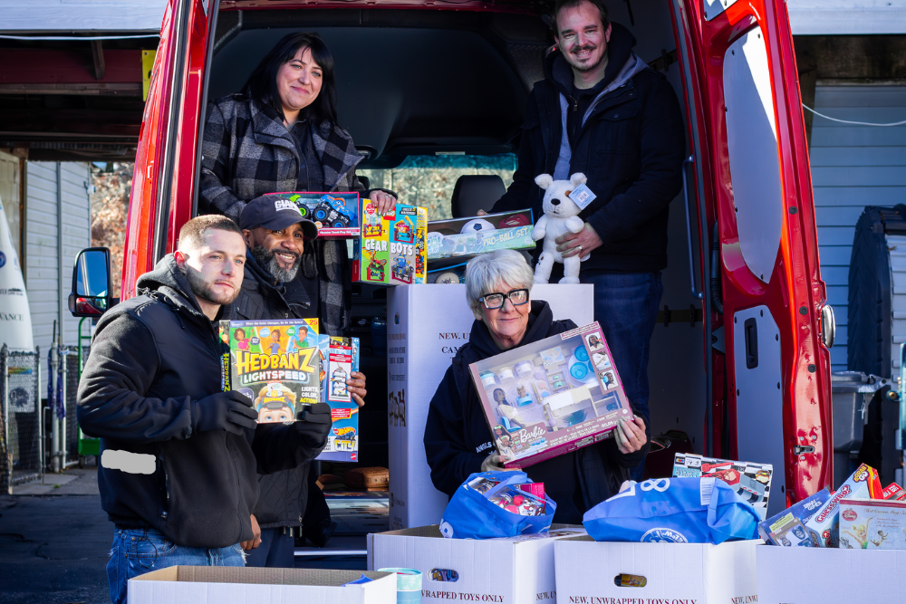 Five people smile while organizing a toy donation from the back of a red van into white boxes.