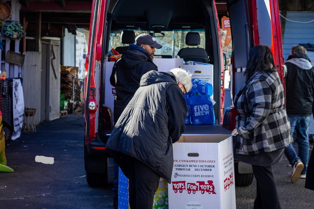 People loading toys into large 'Toys for Tots' collection boxes in the back of a red van
