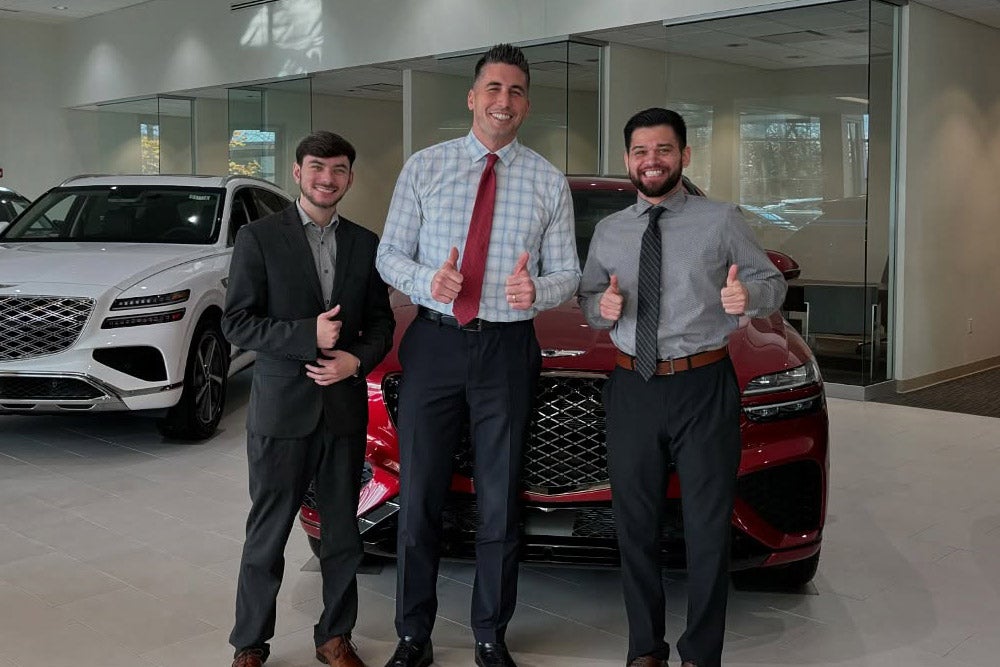 Three car salesmen, dressed in business wear, stand smiling with thumbs up in front of two luxury SUVs.