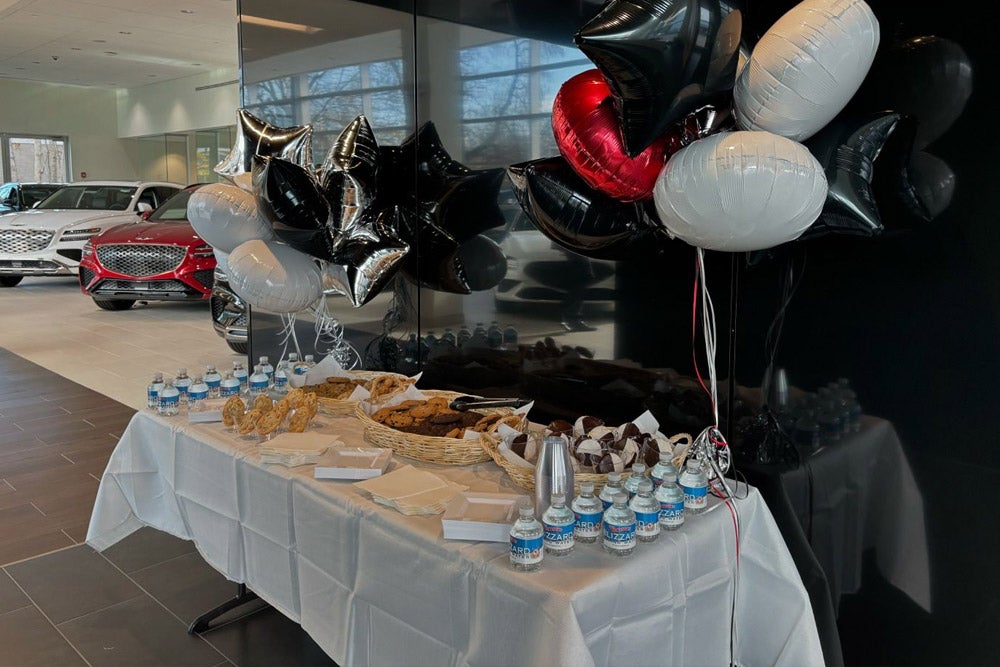 A white-draped table with bottled water and baskets of cookies, muffins, and brownies, with black, white, and red balloons behind it, all set up inside a car dealership.