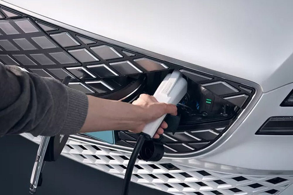 A hand plugging a white charging cable into the front grille of a white electric car.