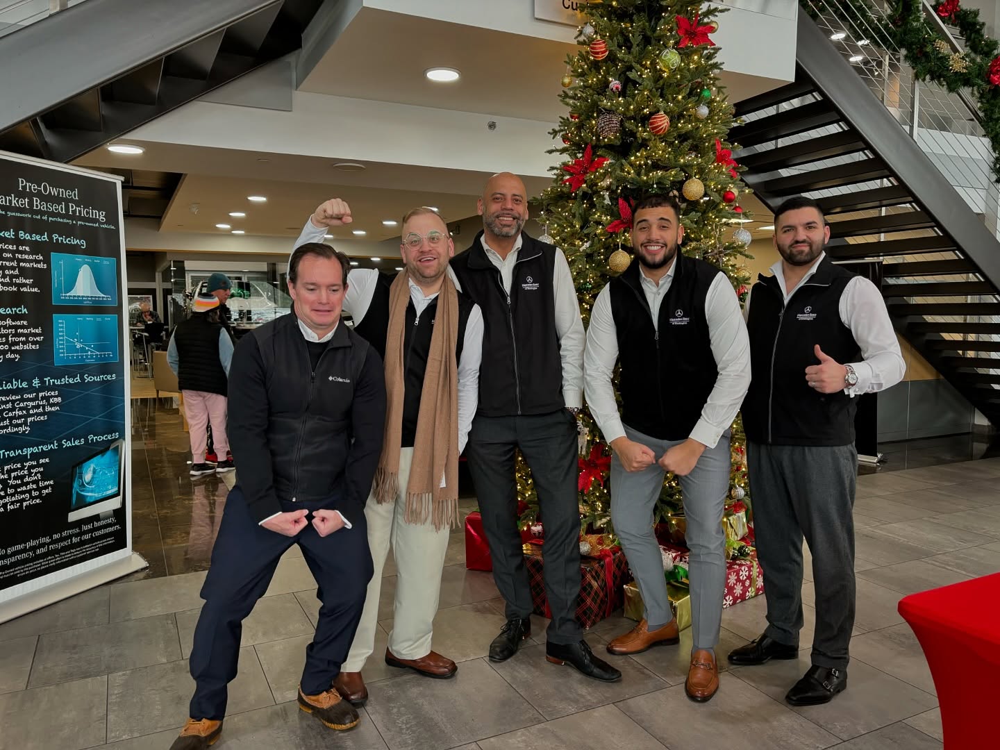 Five men in vests pose playfully in front of a decorated Christmas tree in a modern dealership showroom.