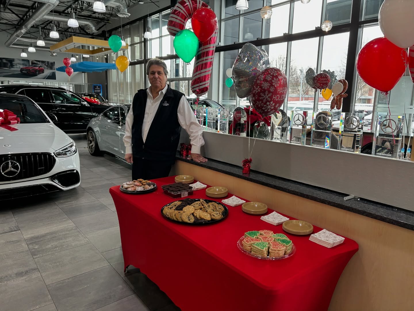 Man in a Mercedes-Benz showroom, standing next to a table with holiday cookies, adorned with festive balloons.