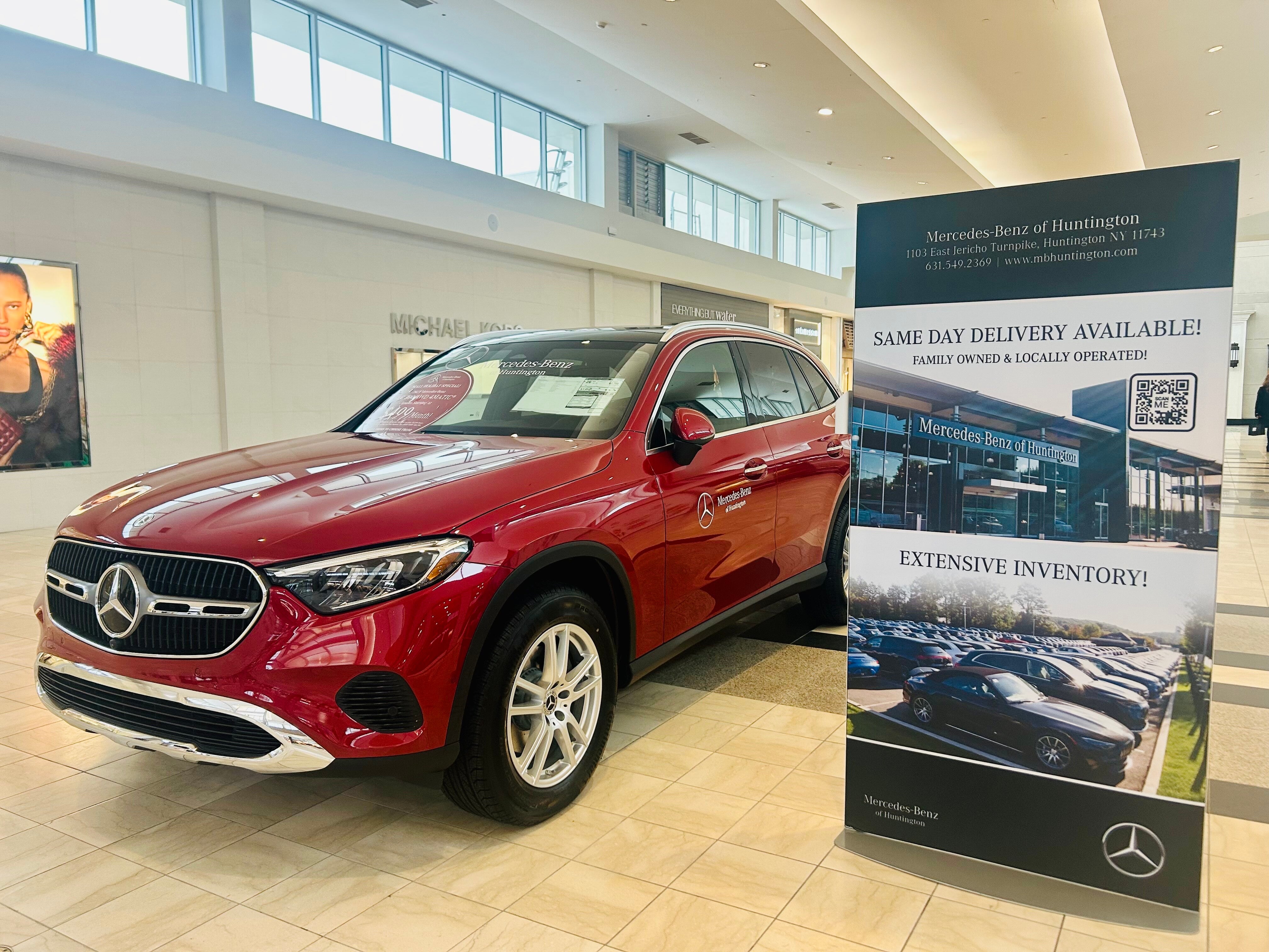A red Mercedes-Benz SUV on display in a mall, with a large 'Mercedes-Benz of Huntington' sign beside it.