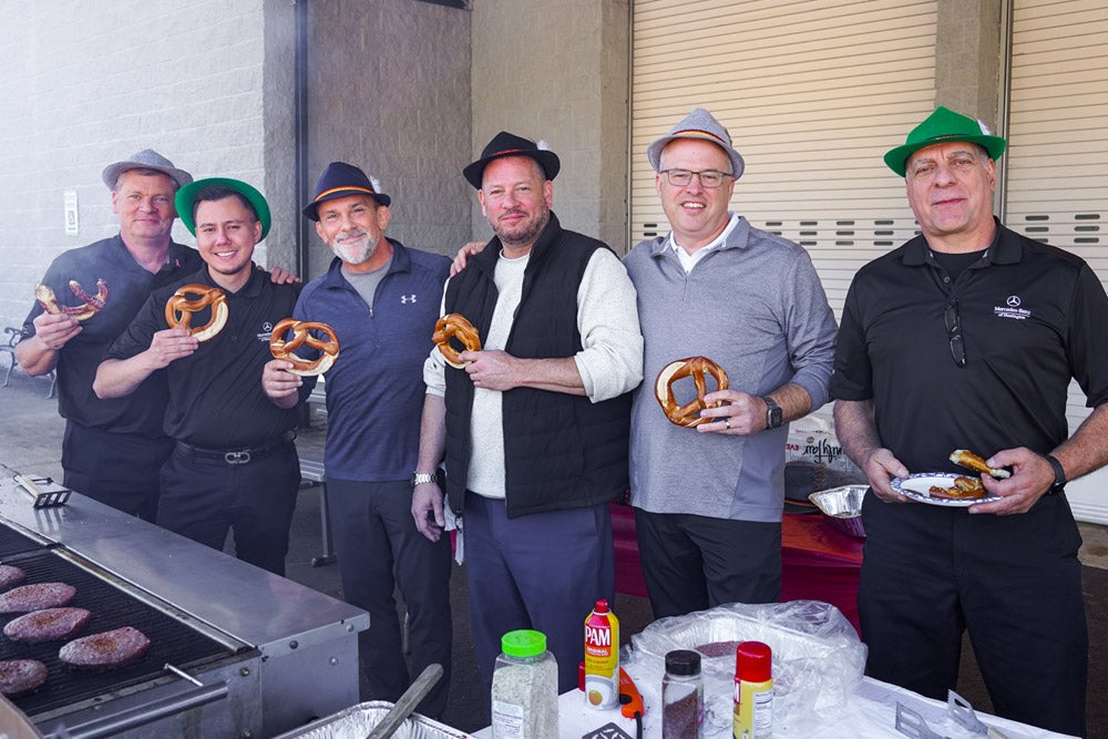 Six men, some wearing traditional German hats, smile and pose with pretzels by a grill cooking hamburgers.