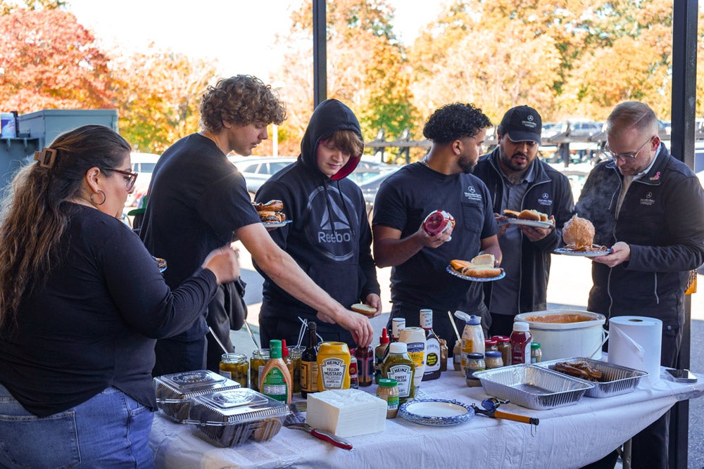 People gathered outdoors around a white table for a casual meal, serving themselves from various condiments and dishes.