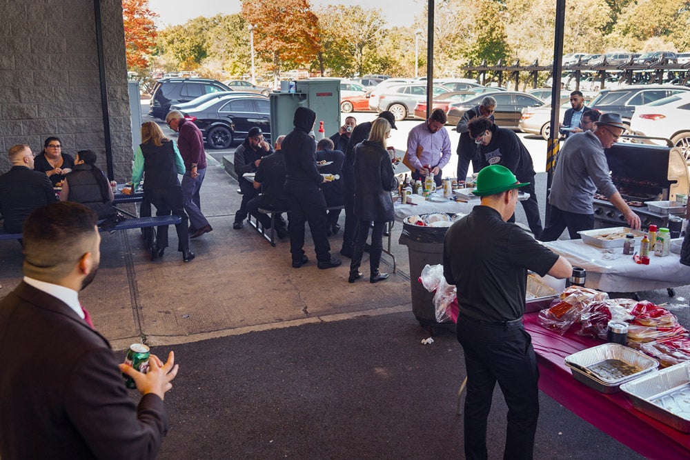 People gather outdoors for a barbecue, with some cooking at a grill and others seated at picnic tables.