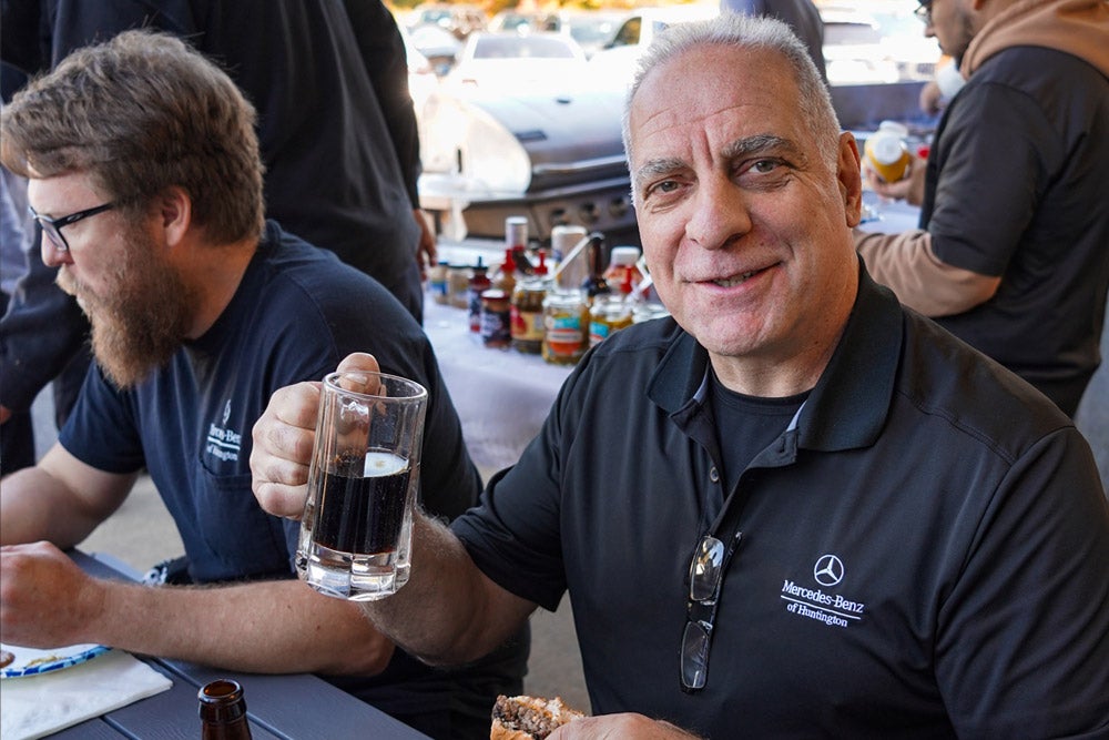 Man in Mercedes-Benz shirt smiling at camera while holding a drink and a burger at an outdoor event.