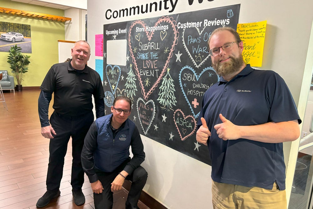 Three Subaru employees in blue shirts stand in front of a community chalkboard about a 'Share the Love Event.'