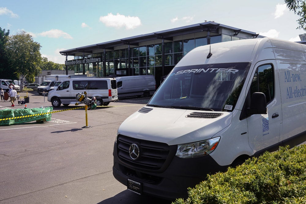 A white Mercedes-Benz Sprinter van, an electric model, is parked outside a Mercedes-Benz dealership.