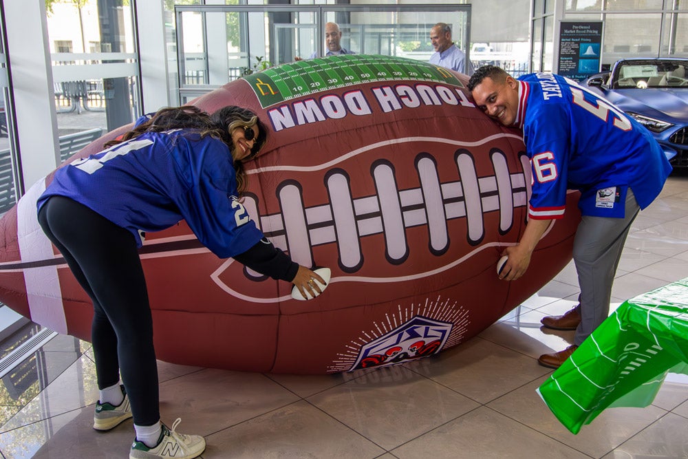 Two smiling people in blue jerseys embrace an oversized inflatable football in a bright room.