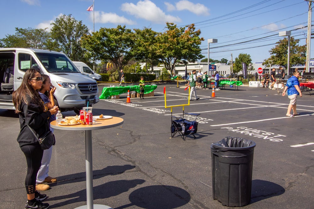 People mingle in a sunny parking lot converted for a tailgate party with food, drinks, and a small football goal.