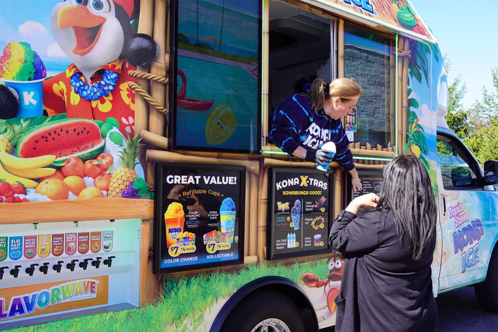 A woman in a 'KONA ICE' hoodie serves shaved ice from a colorful food truck to a customer.