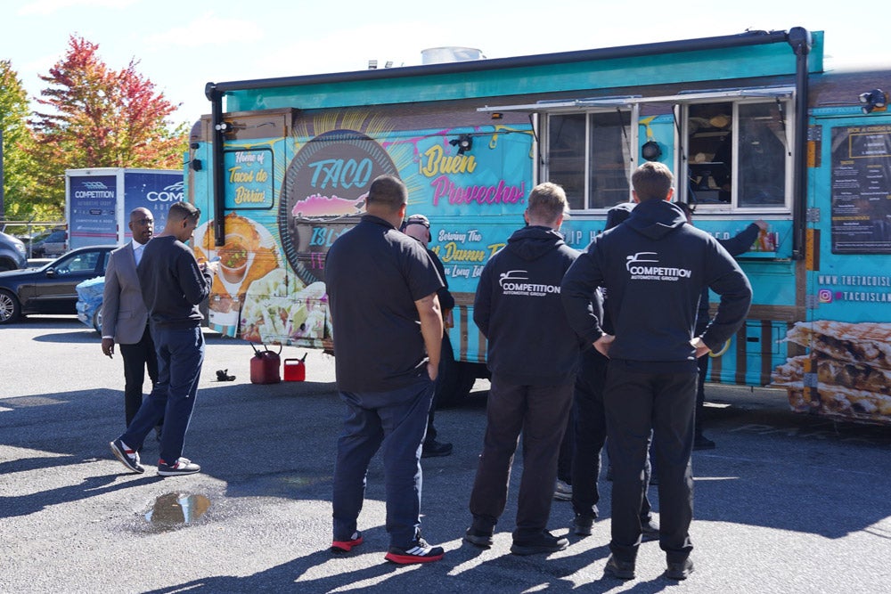 People gather around a turquoise 'Taco Island' food truck under a bright sky with trees in autumn colors.