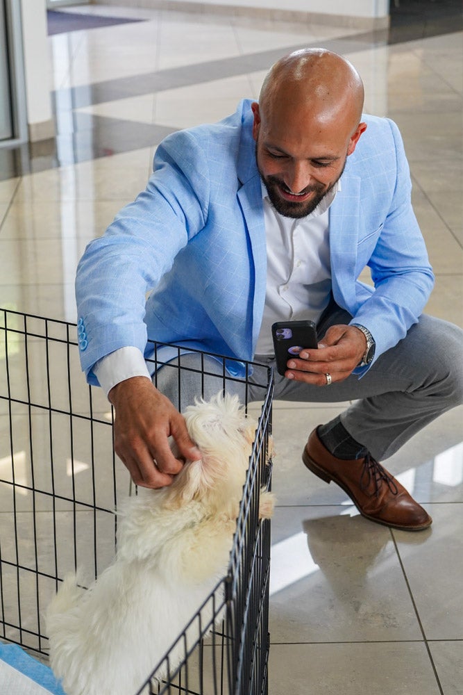 A smiling man in a blue jacket crouches, petting a white puppy in a black wire crate while holding a phone.