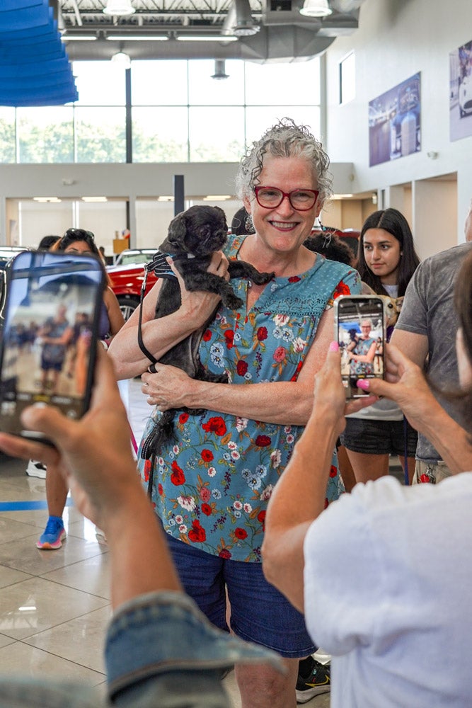 A smiling woman in glasses and a floral shirt holds a small black dog as people photograph them with cell phones.
