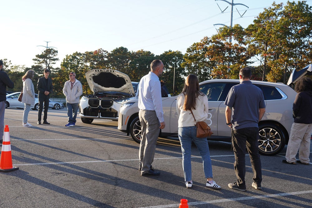People gathered in a sunny parking lot looking at two light-colored SUVs with open hoods and trunks.