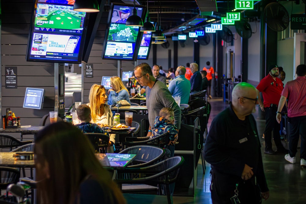 Families and patrons socialize while eating at tables in a lively, modern indoor golf complex with large screens everywhere.