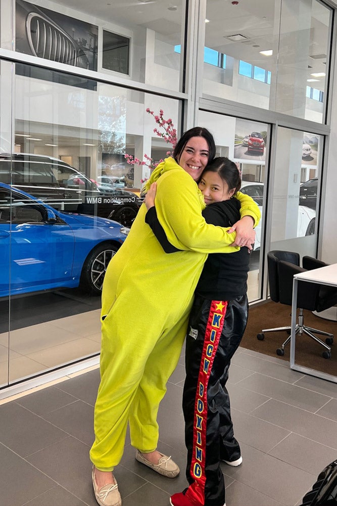A tall woman in a neon green jumpsuit hugs a young girl wearing black kickboxing pants and a black shirt.