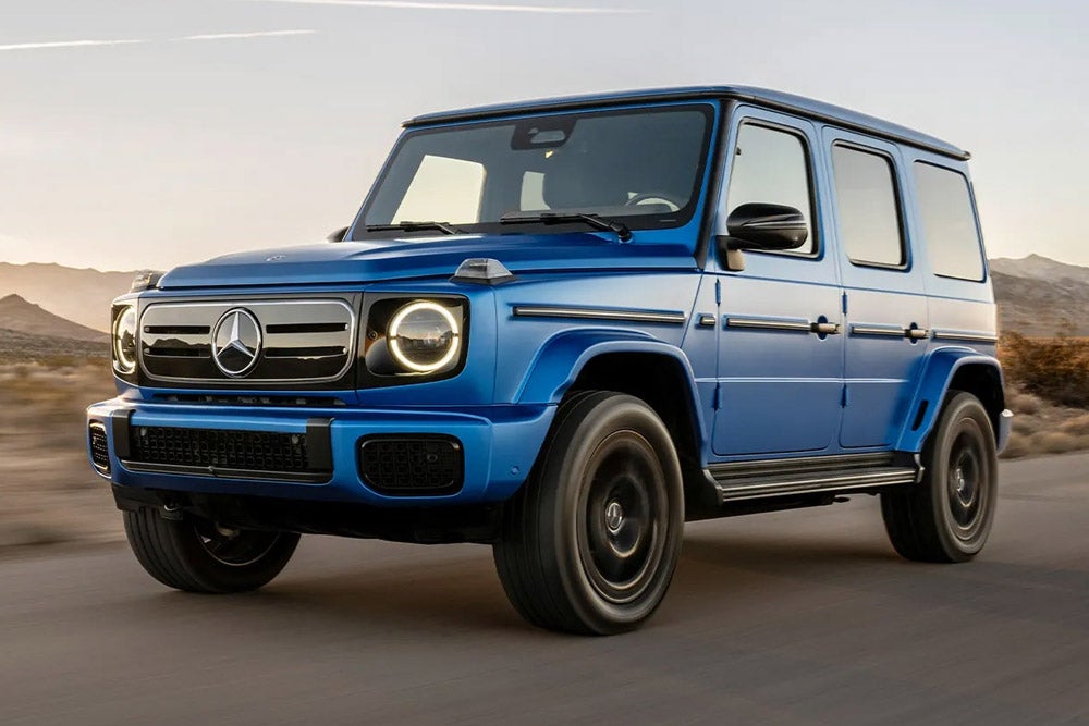 A bright blue Mercedes-Benz G-Class SUV drives down a road, with mountains and a sunset in the background.