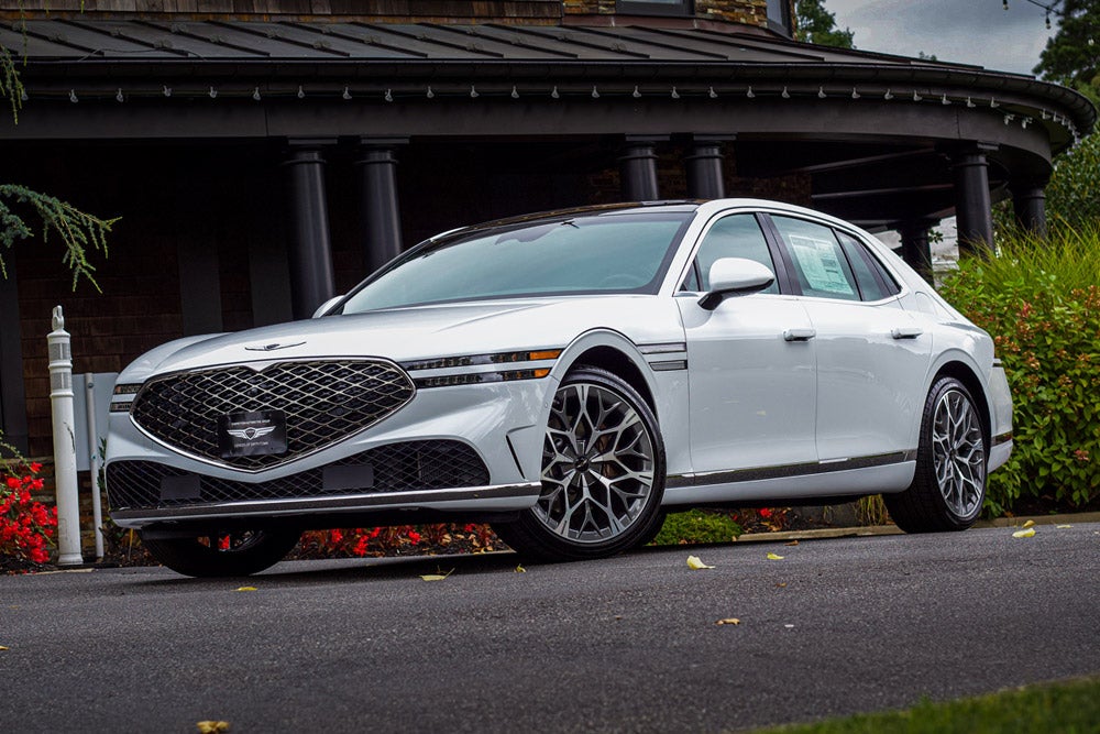 A white Genesis G90 car with a large, diamond-patterned grille parked on asphalt in front of a building.