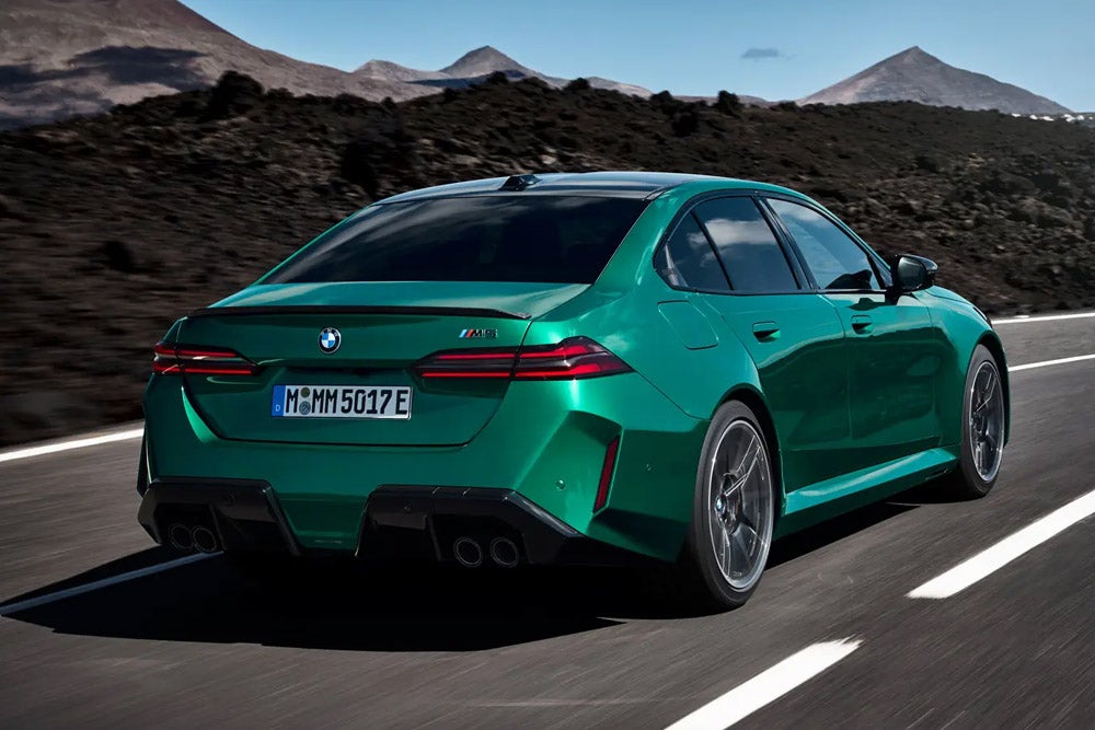 Rear-view of a metallic green BMW sedan driving on a mountain road under a blue sky.