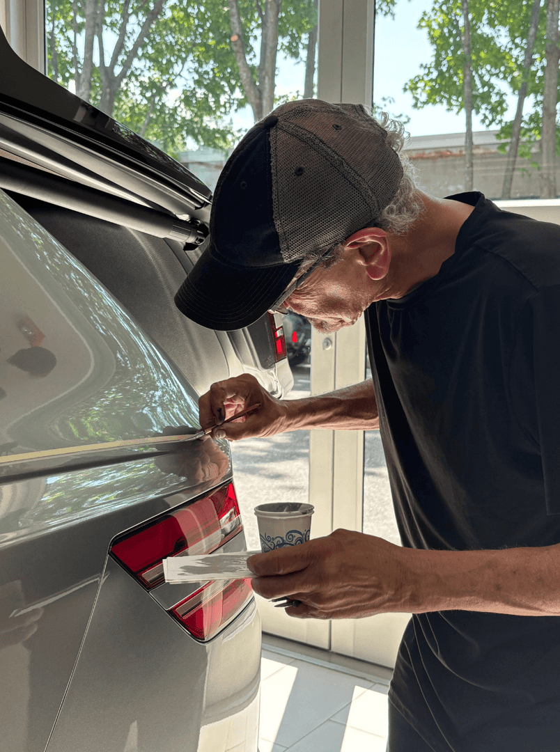 Man in a baseball cap and black shirt meticulously hand-painting a thin stripe on the trunk of a gray car.