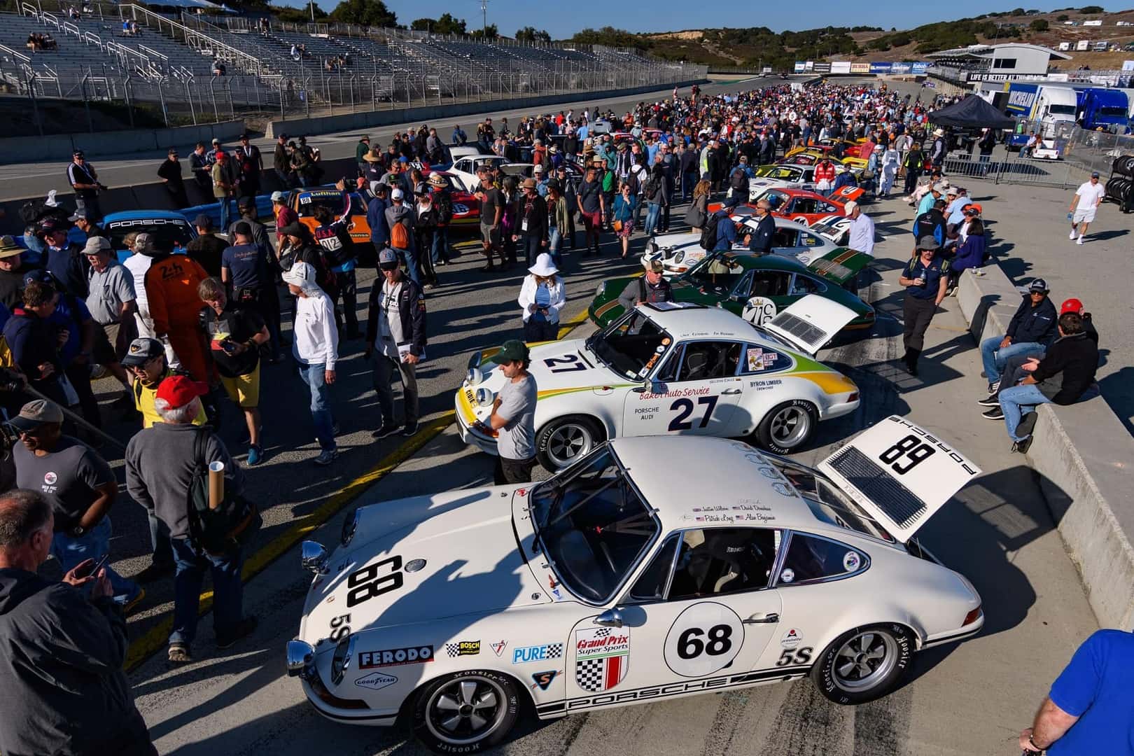 Large crowd around vintage Porsche race cars at a sunny trackside event with empty grandstands in the background.