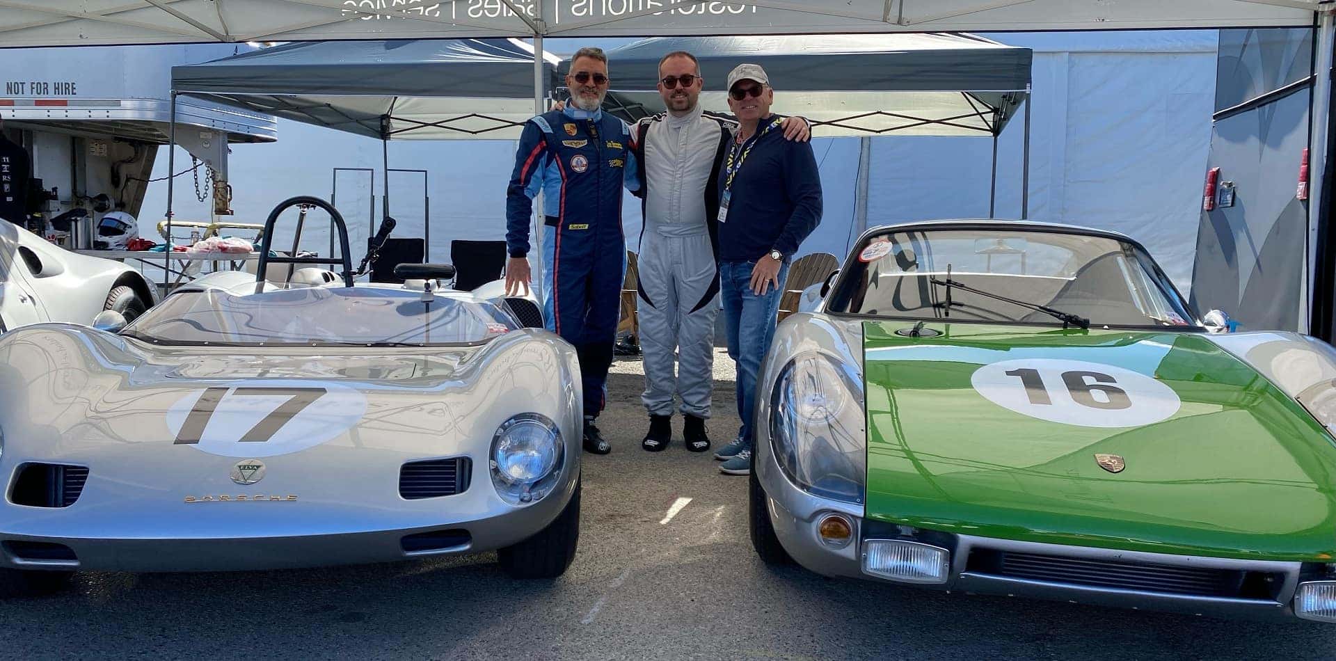 Three men pose between two vintage Porsche race cars, one silver, one green, under a white tent.
