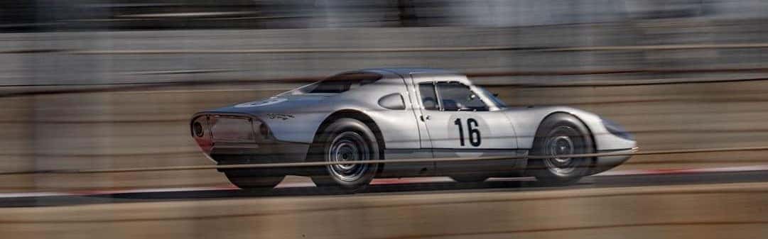 A silver #16 vintage race car speeds right on a track, its background blurred with motion.
