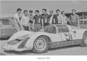 Black and white photo of eight men standing around a #51 Porsche 906 race car at Daytona in 1967.
