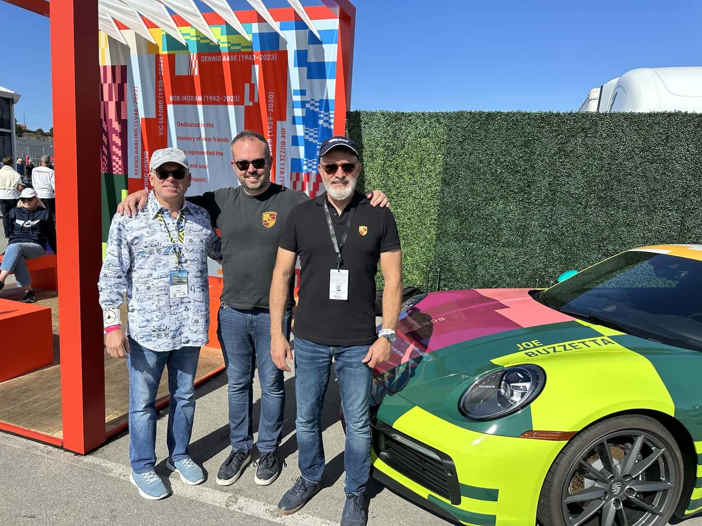 Three men posing with a colorful 'Joe Buzzetta' Porsche car, next to a memorial display.