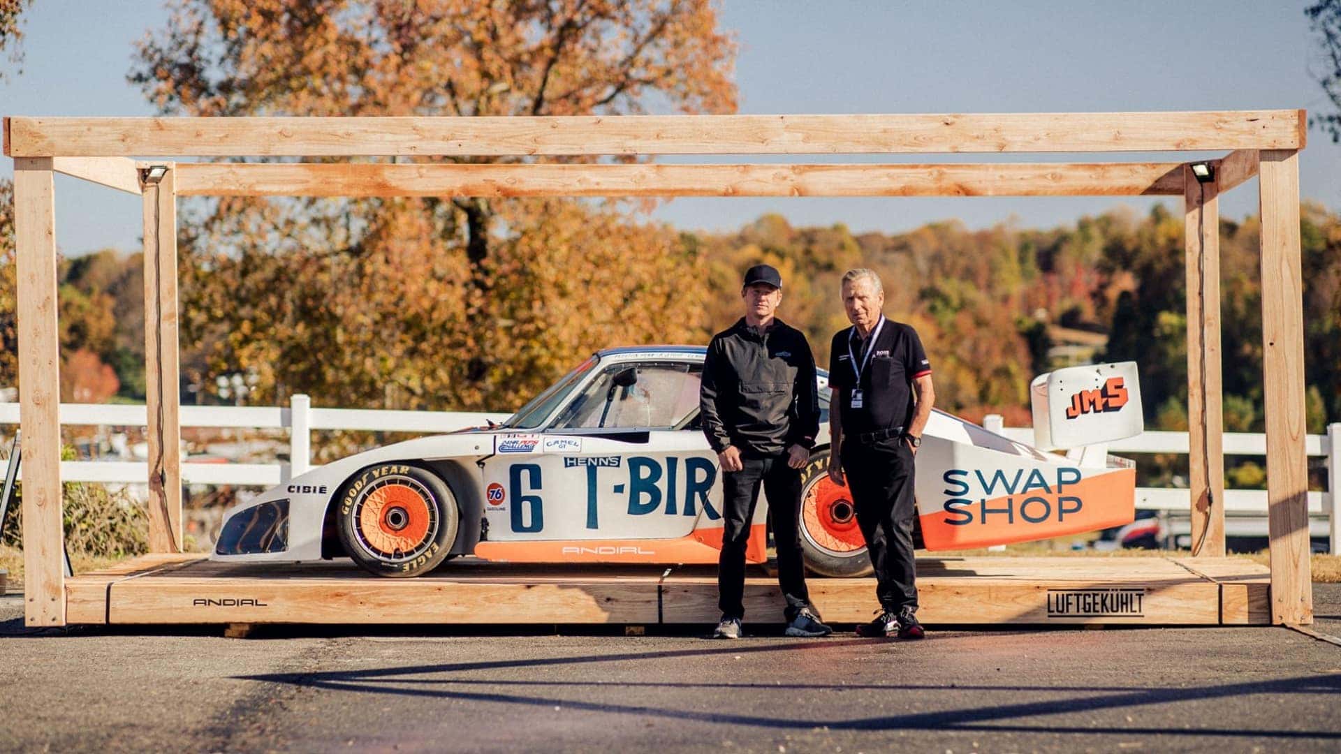Two men stand beside a white and orange racing car with 'T-BIRD' written on its side, inside a wooden frame.