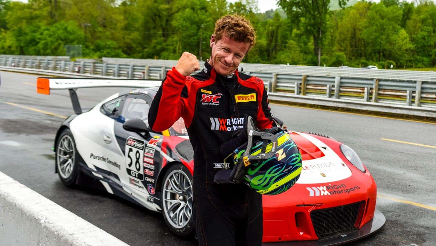 A Patrick Long in a red and black suit with helmet, holding his fist up, next to a #58 Porsche.