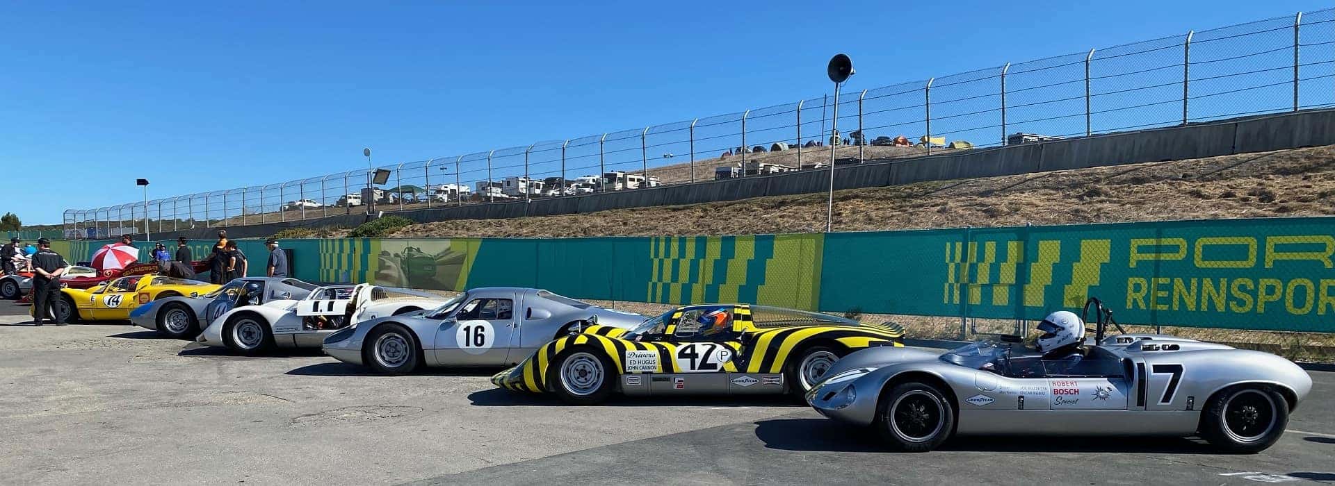 Six vintage race cars, silver and yellow striped, parked on asphalt with spectators and a track fence in the background.