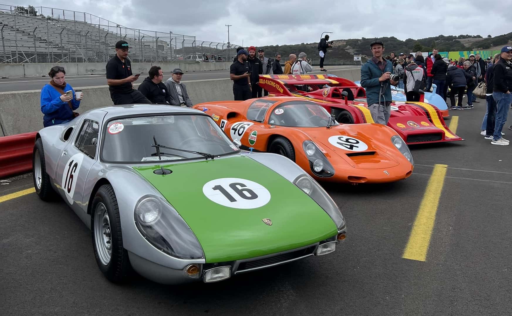 Three vintage race cars parked on a track with spectators in the background, a silver and green car in front.