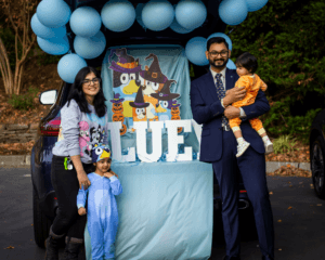 Family in front of a car trunk decorated with Bluey characters and blue balloons for a trunk-or-treat event.