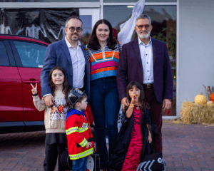 Two men, two women, and two children pose for a photo in front of a red car and a building entrance.