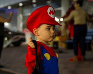Young child dressed as Mario, looking right with a serious expression, holding a black bag.